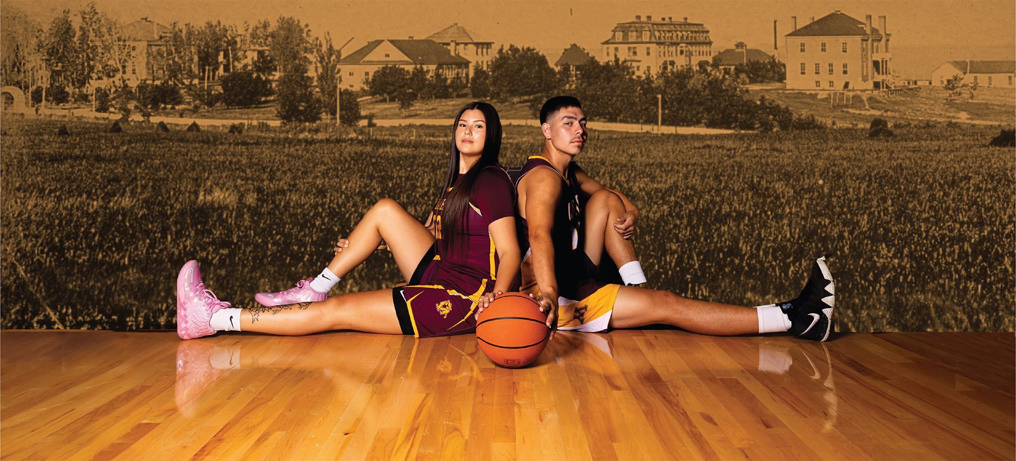 Two basketball players in maroon and gold uniforms sit back-to-back on a polished wooden floor with legs extended, holding a basketball between them; the background shows an old sepia-toned image of buildings and trees.